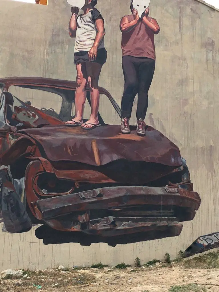 Detalle del mural de gran formato del MIAU de Fanzara: dos figuras con máscaras de gato sobre un coche oxidado destrozado, arte urbano Castellón