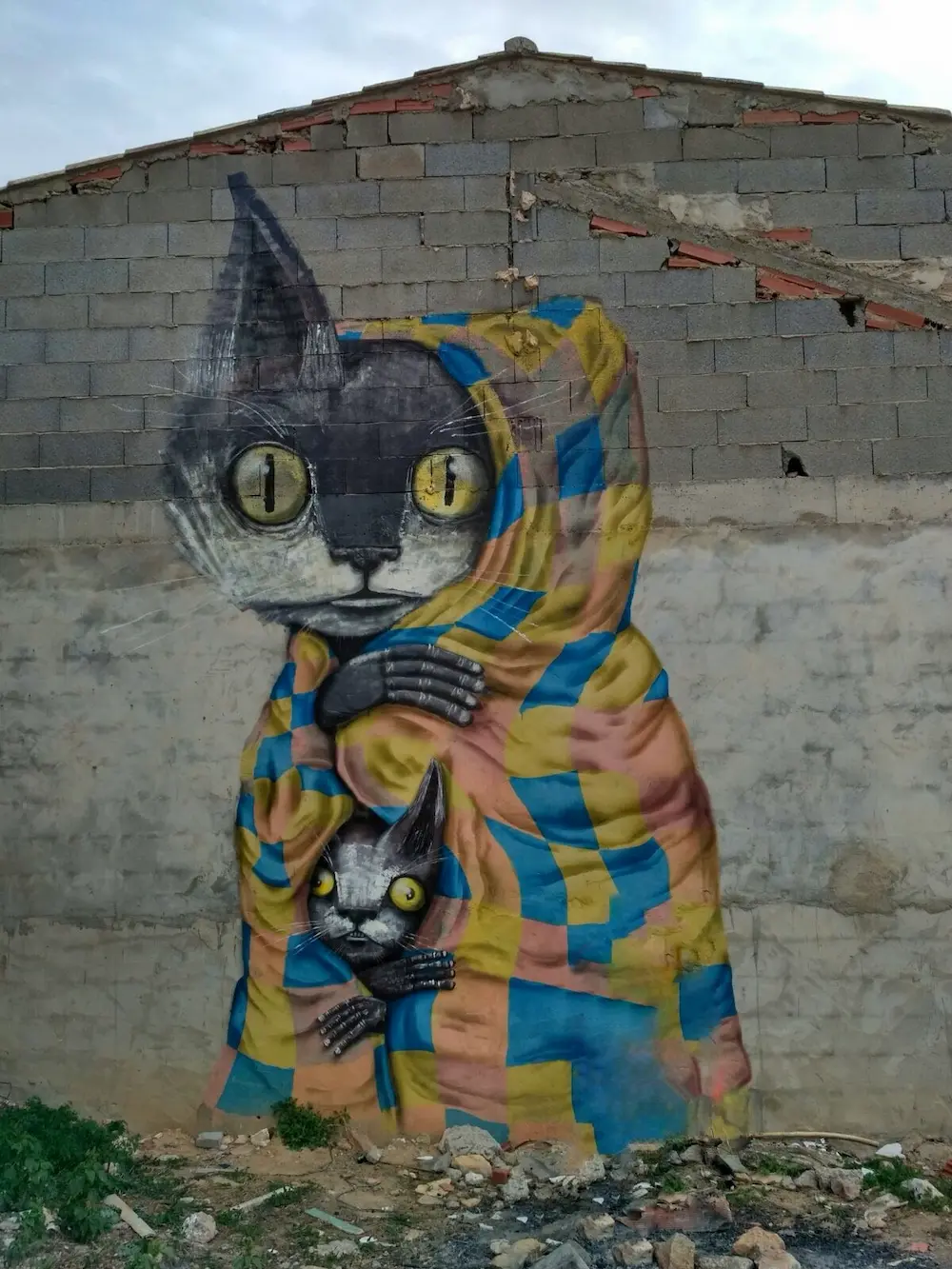 Mural del MIAU de Fanzara: gato gigante con ojos amarillos envuelto en una manta a cuadros azul y amarilla, arte urbano al aire libre en Castellón