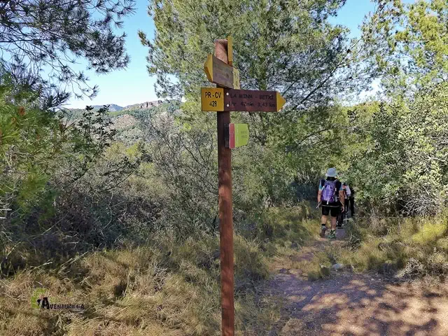 Señal del PR-CV en el sendero de pinar hacia las Peñas Aragonesas, con excursionistas al fondo y el rodeno en el horizonte